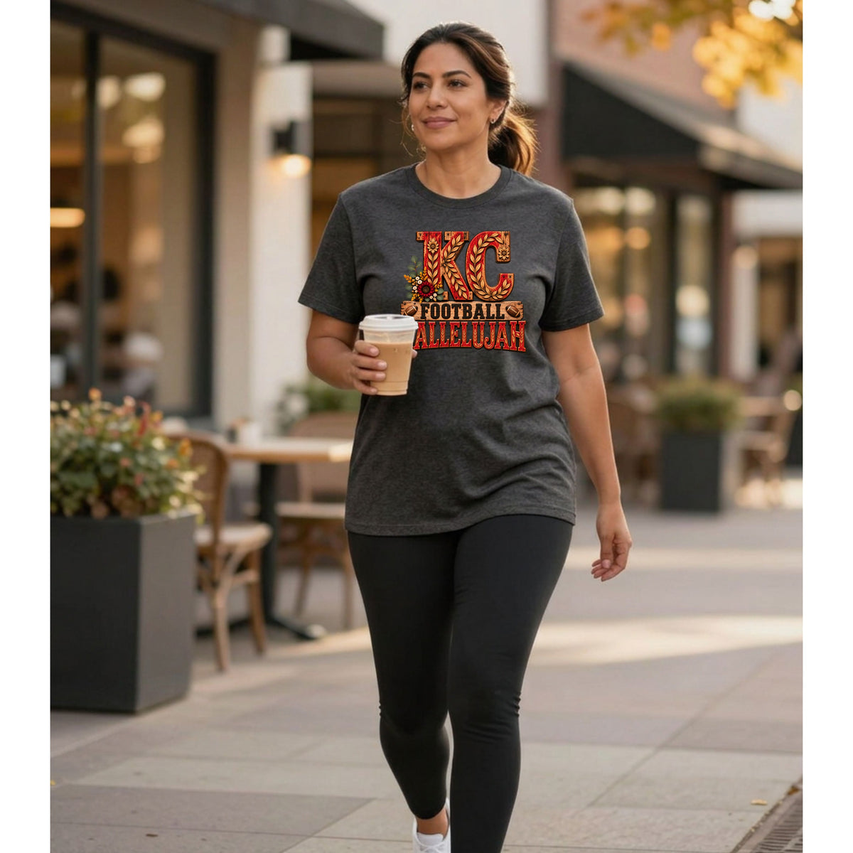 Woman wearing a dark grey t-shirt with "KC FOOTBALL ALLELUJAH" graphic, holding a coffee.
