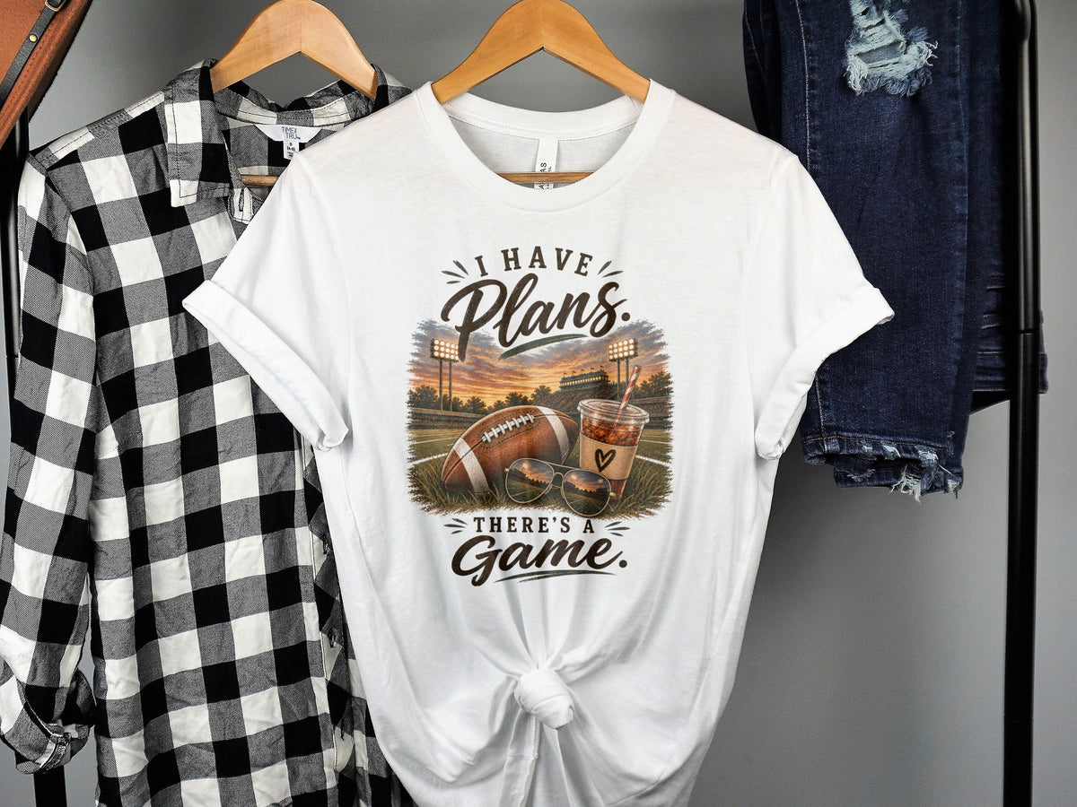 White t-shirt with "I Have Plans" and "There's a Game" text, football, sunglasses, and drink.