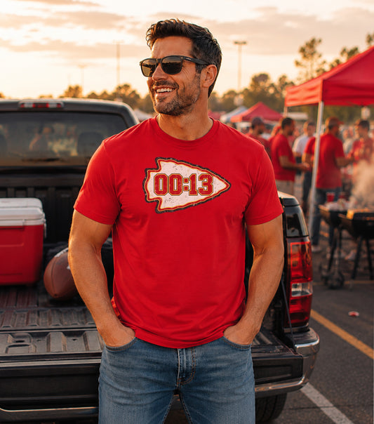 Man in red t-shirt with "00:13" arrowhead logo stands by truck.