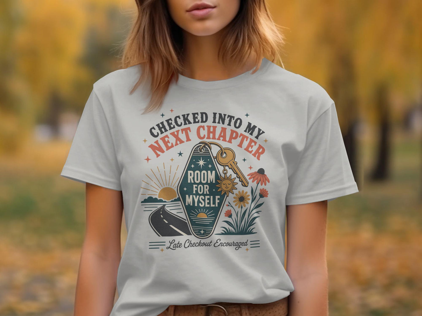 Graphic tee with "Checked Into My Next Chapter Room For Myself" text and sunset, keys, and flowers.